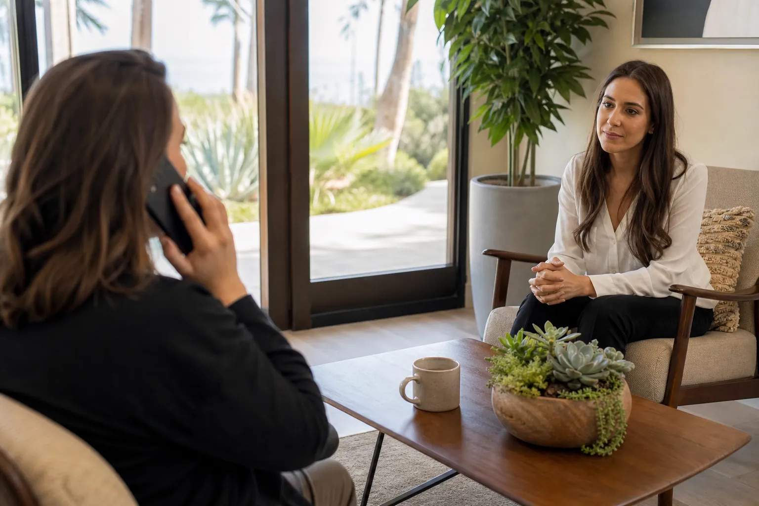 A woman speaks on a phone while another woman listens in a bright, private counseling room with tropical landscaping outside.