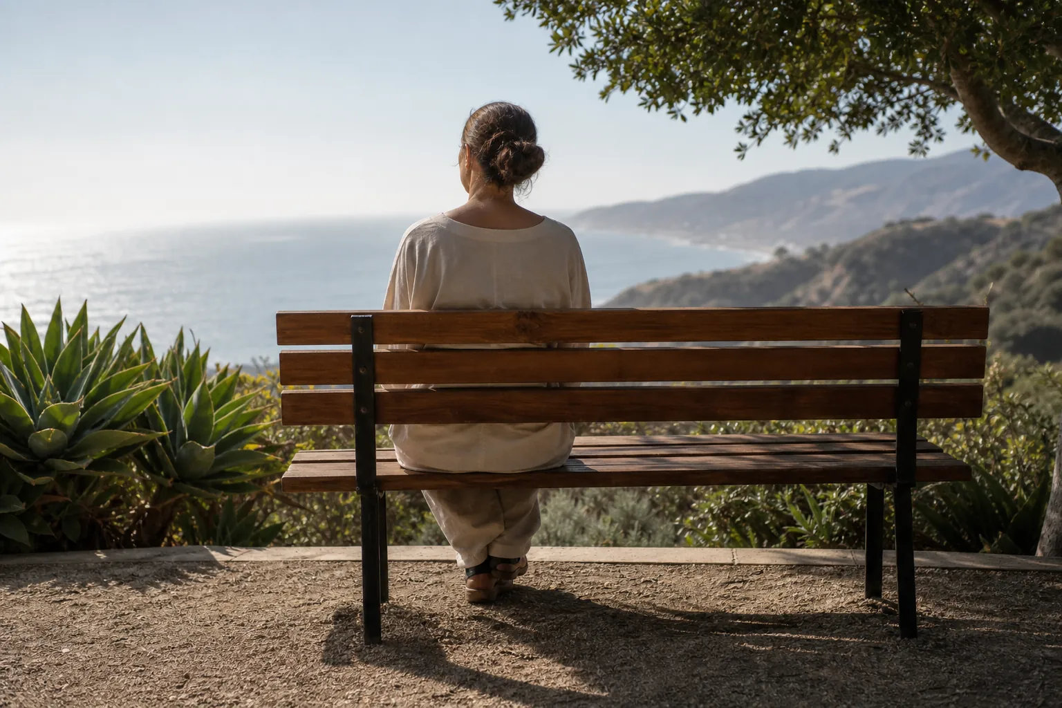 Woman Resting on a Bench Overlooking the Coast A woman sits alone on a wooden bench overlooking the ocean and a mountainous coastline beneath a leafy tree.