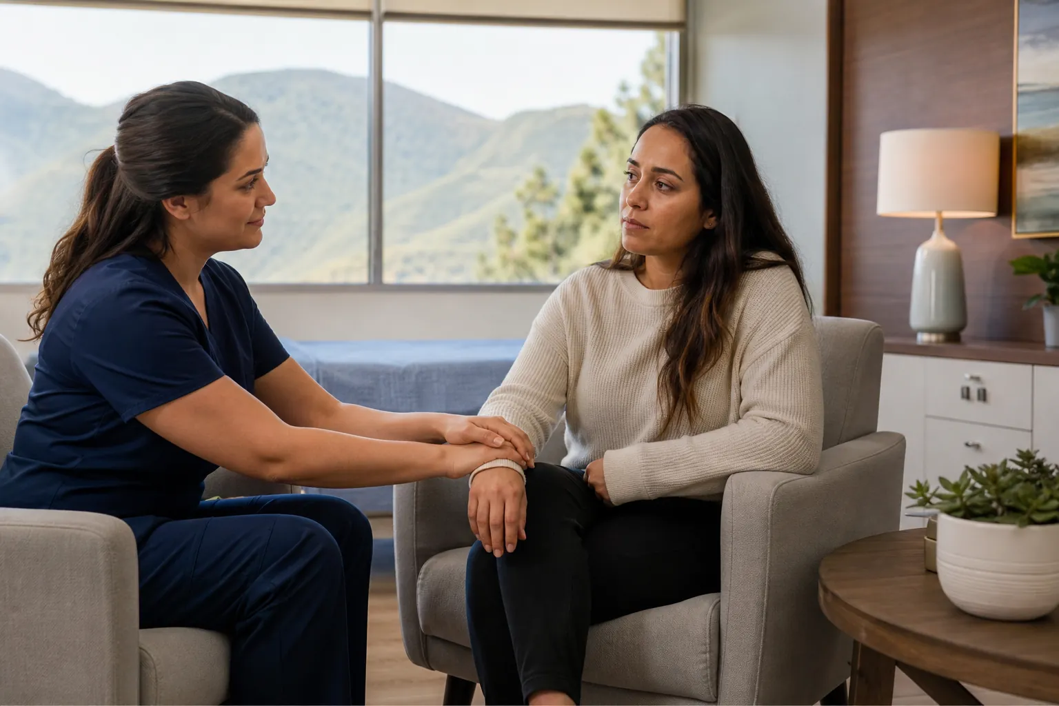 Supportive Conversation With a Healthcare Worker A woman in navy scrubs rests her hands on a seated woman's wrist in a bright room with mountain views.