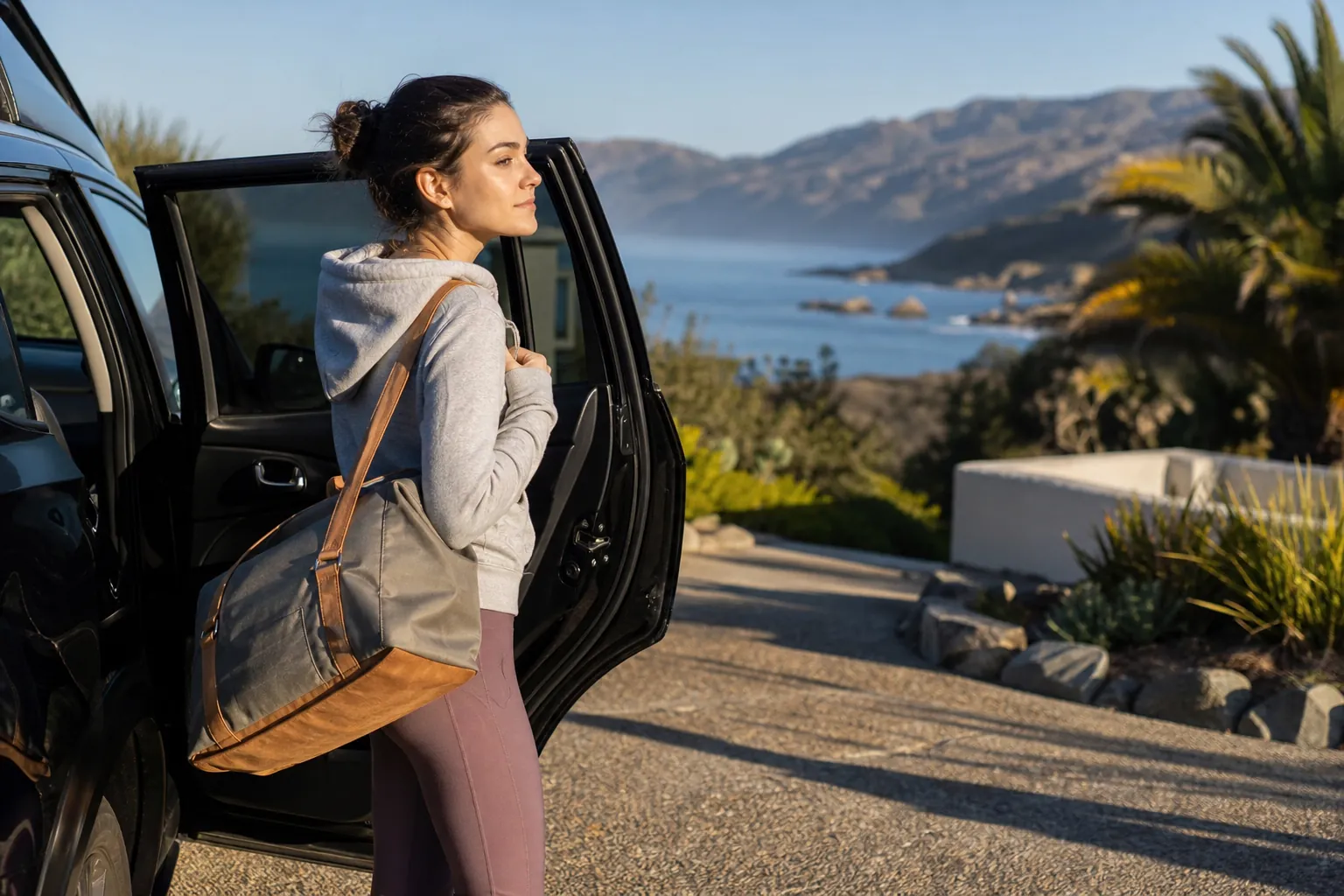 Woman with a duffel bag stands beside an open car door on a driveway overlooking a mountainous coast.