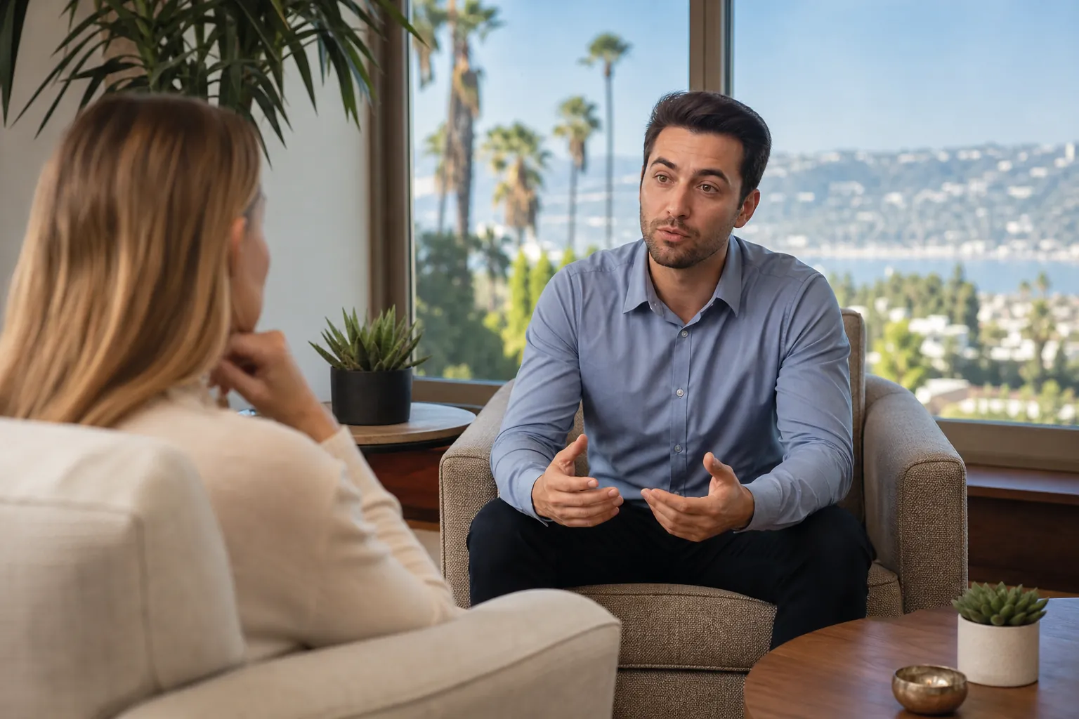A man in a blue shirt gestures while talking with a seated woman in a bright room overlooking palm trees and water.