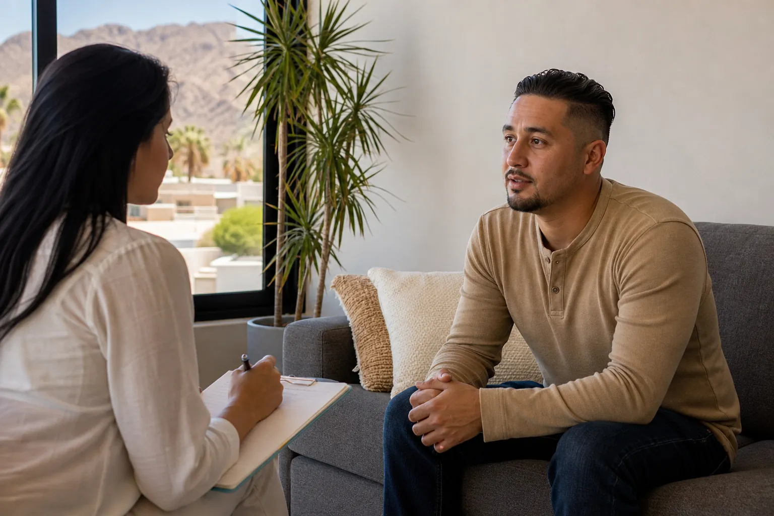 A man seated on a gray sofa talks with a woman holding a clipboard, with desert mountains visible through the window.