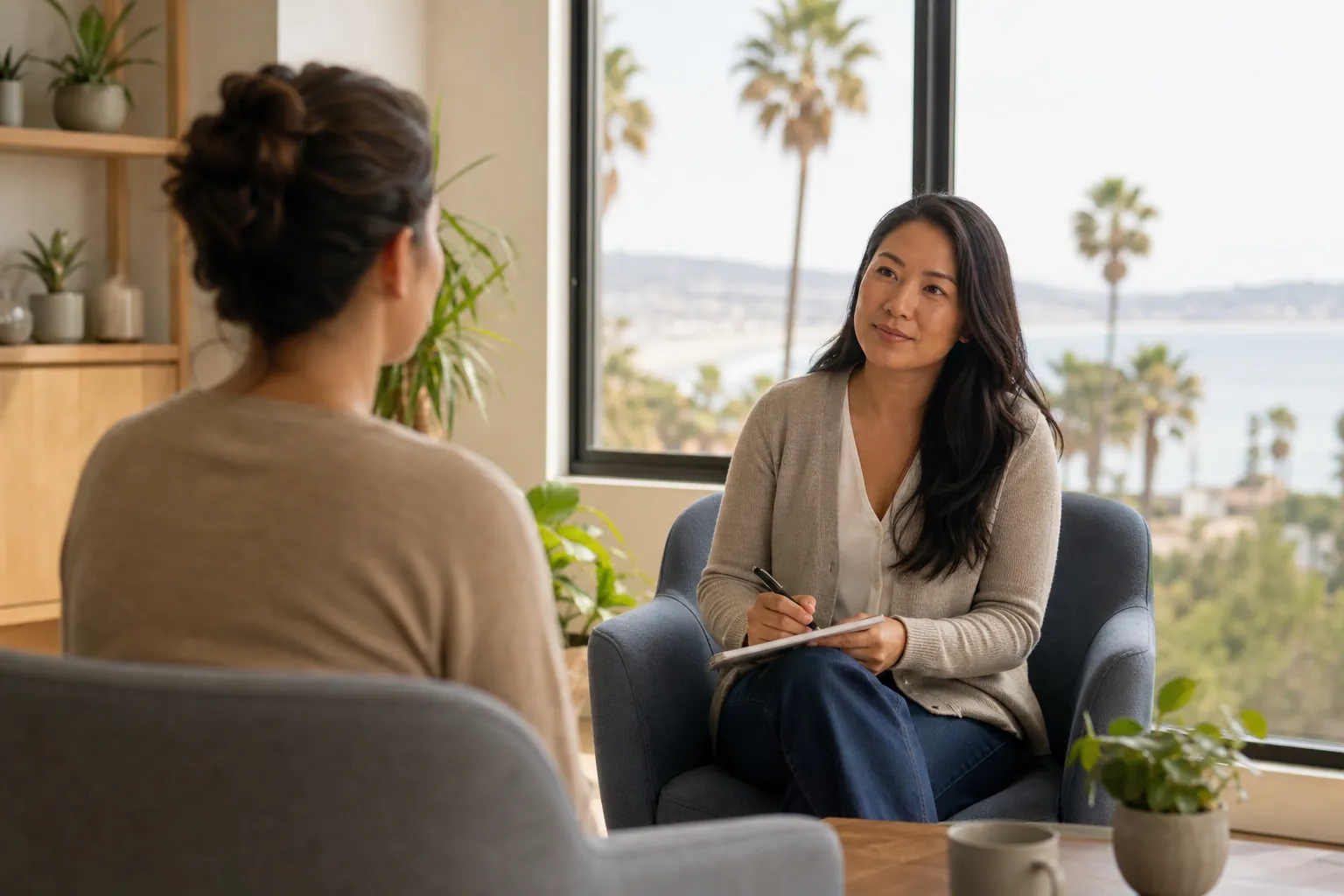 Two women sit facing each other in a bright office, one holding a pen and clipboard, with palm trees visible outside.