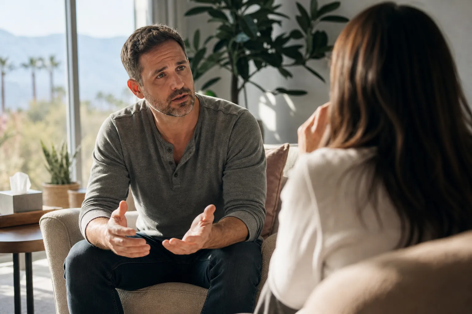A seated man gestures while talking with a woman in a bright room with plants and desert mountains visible through windows.