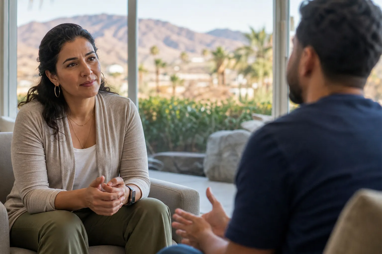 A woman listens to a man gesturing from a facing chair, with desert mountains and palm trees visible through large windows.
